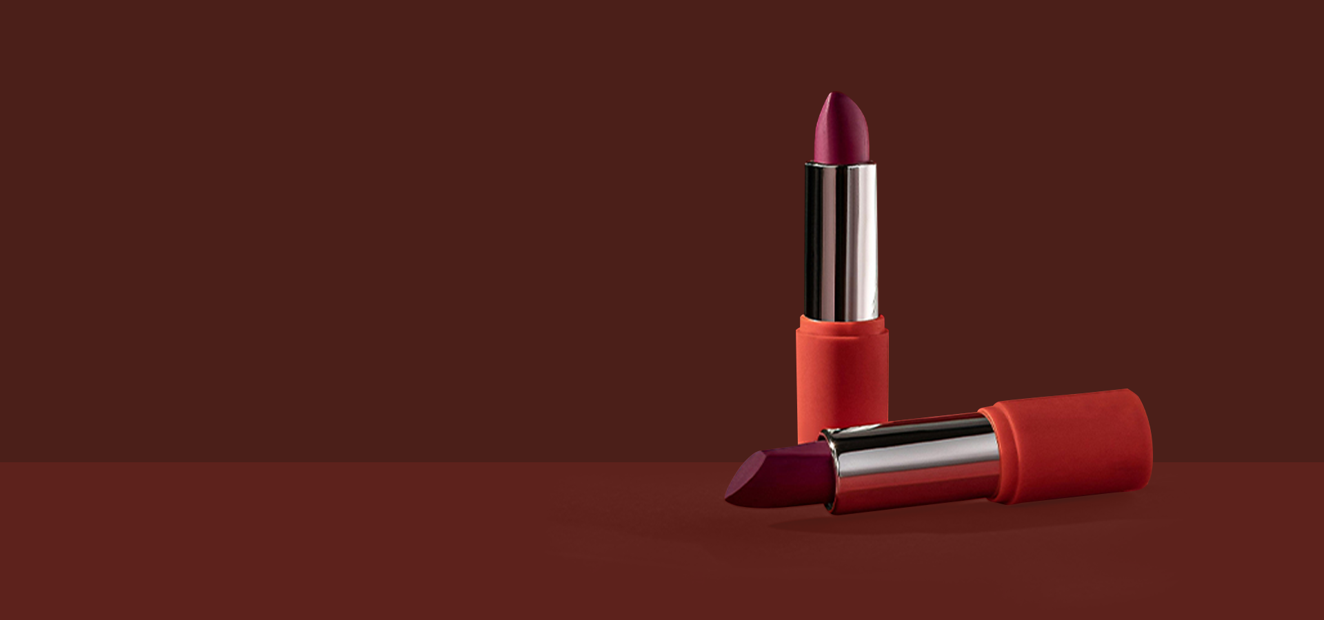 Two lipsticks in red and orange cases on a dark brown background, one standing upright and one lying on its side showing the lipstick bullet.